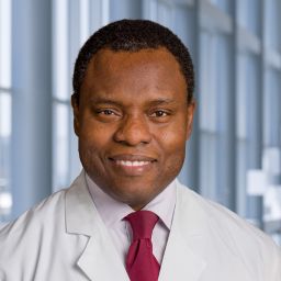 Dr. Tayo Addo wearingi a white shirt with red necktie and a white lab coat standing in the Clements University Hosptial lobby