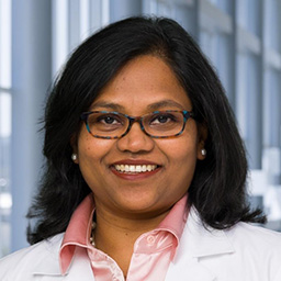 Dr. Vidushi Golla wearing glasses, a pink shirt, and a white lab coat standing in the Clements University Hospital lobby