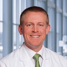 Dr. Kyle Westbrook wearing a white shirt with a green tie and a white lab coat standing in the Clements University Hospital lobby