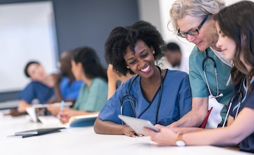 classroom of medical staff seated at tables; 3 women in blue scrubs in foreground look at document held by woman in center